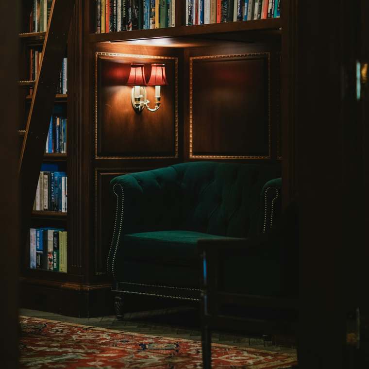 A green velvet armchair beneath a lamp in a private library, a ladder against the shelves